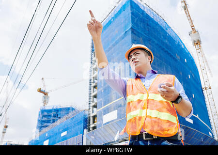 Stirnrunzelnd vietnamesischen Auftragnehmer in hellen orange Weste an Gebäude auf der Baustelle zeigen Stockfoto