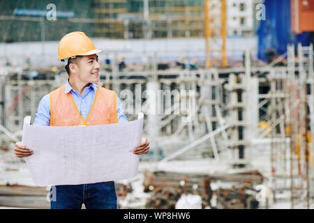 Positive professionelle Asiatische Bauingenieur mit Gebäude Blaupause in seine Hände lächelnd und Wegsehen Stockfoto