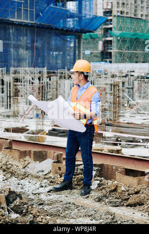 Positive asiatischen Ingenieur tragen leuchtend orange Weste und Helm, wenn auf Neues Projekt auf der Baustelle arbeiten Stockfoto