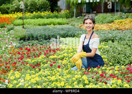 Glückliche junge Frau in Schürze und Gummi finster blicken genießen Sie die Zeit in Ihrem Garten mit vielen schönen blühenden Blumen Stockfoto
