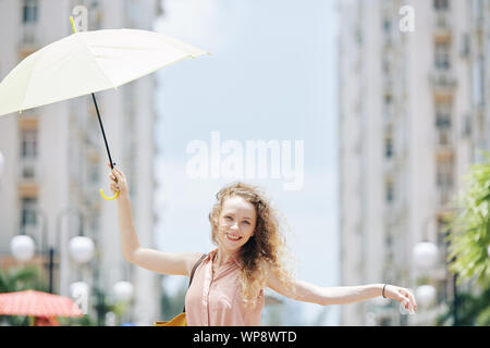 Glückliche junge hübsche Frau, die zu Fuß auf der Straße mit Regenschirm in der Hand auf windigen Tag Stockfoto