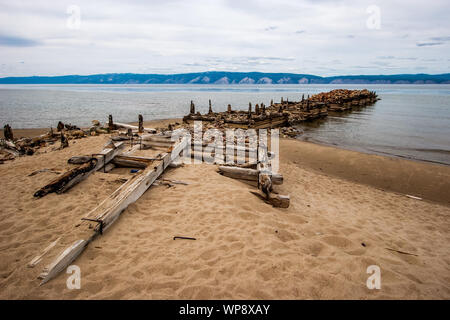 Alte kaputte Boot Pier auf der Insel Olchon am Baikalsee. Morsche Balken und Protokolle. Am Ufer von Steinen und Sand. Hinter dem Bergsee. Stockfoto