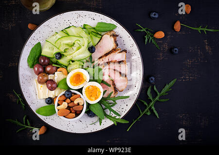 Ketogenic Diät. Keto Brunch. Gekochte Eier, Schweinesteak und Oliven, Gurken, Spinat, Brie Käse, Nüsse und Heidelbeeren. Ansicht von oben Stockfoto