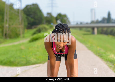 Junge athletische afrikanisches Mädchen eine Pause vom Joggen nach vorn auf die Knie gelehnt verspannt ruht während der Wiedergabe von Musik über Ohrstöpsel Stockfoto