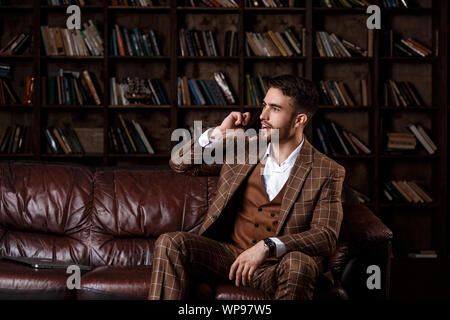 Junge Bartgeier Geschäftsmann in einem braunen Anzug im Gespräch am Telefon mit einem Lächeln auf seinem Gesicht, während in der Bibliothek Büro zu sitzen. Stockfoto