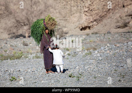Frau trägt an Kopf große Scheiben von Heu und halten ein Kind in der ariden Landschaft im Oman Stockfoto