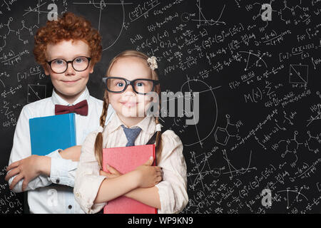 Smart Kids portrait. Kleine Mädchen und jungen Studenten holding Bücher und stehend gegen Blackboard Hintergrund mit Wissenschaft und Mathematik Formeln Stockfoto