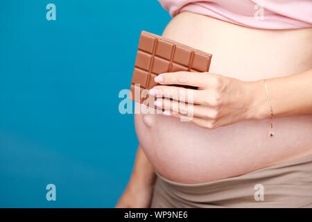 Studio geschossen von jungen schwangeren Frau mit Schokolade in der Nähe von ihrem Bauch vor blauem Hintergrund - Schwangerschaft und Hormone Konzept Stockfoto