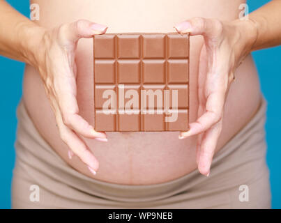 Studio geschossen von jungen schwangeren Frau mit Schokolade in der Nähe von ihrem Bauch vor blauem Hintergrund - Schwangerschaft und Hormone Konzept Stockfoto