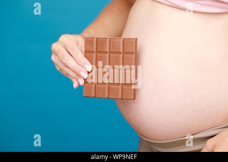 Studio geschossen von jungen schwangeren Frau mit Schokolade in der Nähe von ihrem Bauch vor blauem Hintergrund - Schwangerschaft und Hormone Konzept Stockfoto