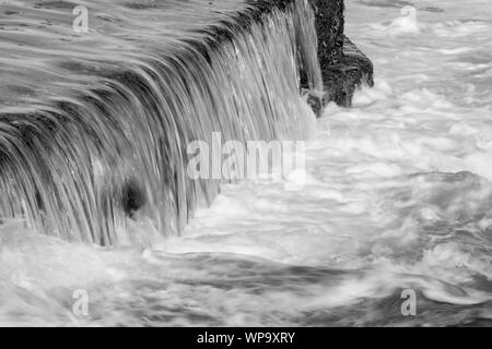 Schwarze und Weiße abstrakte Fotografien einer Meereslandschaft mit starken Rückspülen mit Wasser fließt über eine Flutwelle pool Wand auf einem niedrigen Verschlusszeit - leistungsstark Stockfoto