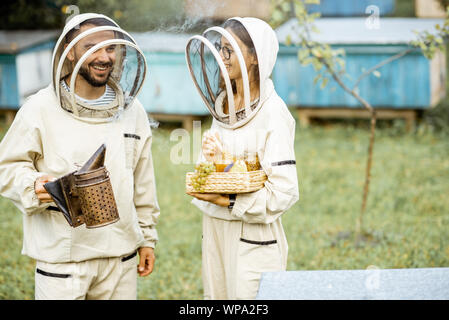 Zwei beekepers in Schützende Uniform stehen zusammen mit beesmoker und Honig, schmecken frische Ware auf dem Bienenhaus Stockfoto