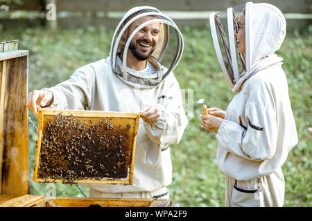 Zwei junge beekepers in Schützende Uniform arbeiten auf einer kleinen Imkerei Bauernhof, Wabe, vom hölzernen Bienenstock Stockfoto