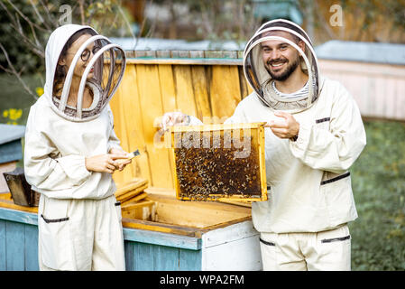 Zwei junge beekepers in Schützende Uniform arbeiten auf einer kleinen Imkerei Bauernhof, Waben aus dem Holz- Bienenstock Stockfoto