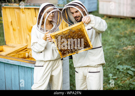 Zwei junge beekepers in Schützende Uniform arbeiten auf einer kleinen Imkerei Bauernhof, Waben aus dem Holz- Bienenstock Stockfoto