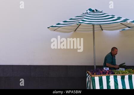 Funchal, Madeira, Portugal - 16 August 2019: Ein Mann verkauft Obst und Gemüse, das unter einem gestreiften Schirm auf der Straße Stockfoto