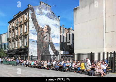 Touristen auf die Sitzgelegenheiten im Freien an der Hootenanny pub Sitzen im Bereich namens "Billy" Biergarten nach dem Wandbild von Billy Connolly, der berühmte Stockfoto