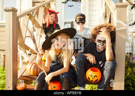 Multi-ethnischen Gruppe von Kindern tragen Halloween Kostüme bei Kamera schaut beim Sitzen auf der Treppe der eingerichteten Haus in Trick oder Festlichkeit Saison Stockfoto