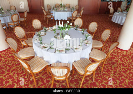 Runde Tische, mit einem weißen Tuch bedeckt, serviert mit grünen Gläsern und verziert mit Blumen, stehen auf einem bunten Teppich. Stockfoto