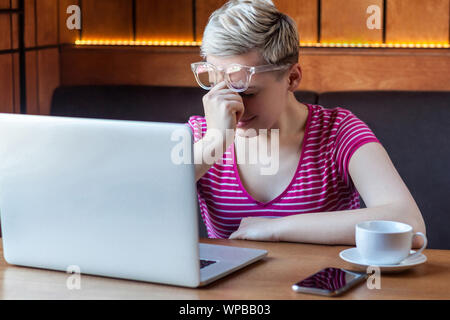 Seitenansicht Portrait von unglücklichen jungen Mädchen mit blonden kurzen Haaren in Rosa t-shirt und Brillen im Cafe sitzen und Arbeiten am Laptop, geschlossenen Augen ein Stockfoto