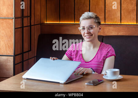 Porträt der schönen Zufriedene glückliche junge bussineswoman mit blonden kurzen Haaren in Rosa t-shirt sitzt im Cafe, Nacharbeiten und Cam Stockfoto