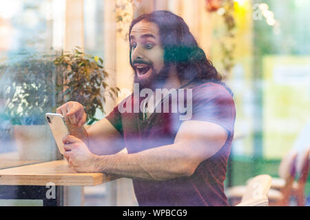 Seitenansicht Portrait durch das Fenster der aufgeregten hübscher junger Erwachsener Sportler Mann mit lockigem Haar lang sitzen auf Cafe, Telefon mit überrascht Fa Stockfoto