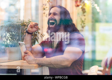 Seitenansicht Portrait durch das Fenster von zufriedenen hübscher junger Erwachsener sportliche Meister Mann mit lockigem Haar lang sitzen auf Cafe, Telefon und tr Stockfoto