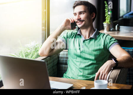 Junge glücklich Geschäftsmann in grünem t-shirt am Schreibtisch mit Laptop sitzen, am Telefon sprechen und Suchen außerhalb und toothy Lächeln. Unternehmen und freiberuflich Stockfoto