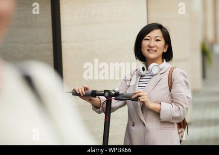 Taille bis Portrait von jungen asiatischen Frau, Elektroroller und Suchen an der Kamera, während draußen in der Stadt Straße Posing, kopieren Raum Stockfoto