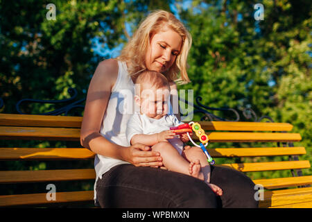 Baby Mädchen mit Spielzeug spielen Sitzen auf den Knien ihrer Mutter im Sommer Park. Familie Spaß im Freien sitzt auf der Bank Stockfoto