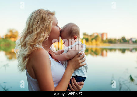 Junge Mutter halten und küssen Baby Girl von Sommer River. Frau mit Kind bei Sonnenuntergang. Familie verbringt die Zeit zusammen Stockfoto