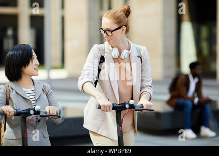 Porträt von zwei zeitgenössische junge Frauen reiten Elektroroller in Street, Fokus auf lächelnde rothaarigen Mädchen im Gespräch mit Freund, Kopie Raum Stockfoto