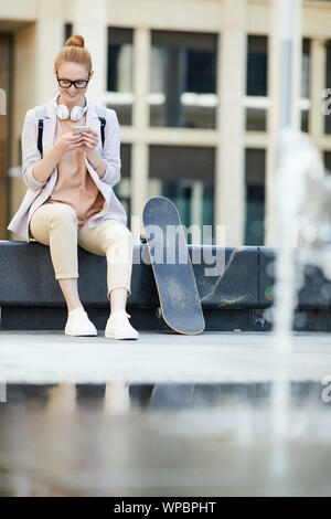 In voller Länge Porträt des zeitgenössischen junge Frau mit Smartphone und glücklich lächelnd, während sie auf der Bank im städtischen Raum einstellen, kopieren Stockfoto