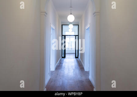 Eine große Halle in einem hohen Raum mit weißen Wänden, drei Kronleuchtern sichtbar und eine offene Tür am Ende des Ganges beleuchtet vom Licht her Stockfoto