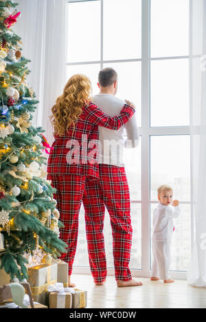 Happy Family im karierten Schlafanzug in der Nähe der großen Fenster im Wohnzimmer mit einem Weihnachtsbaum. Mutter, Vater und Sohn Warten auf den Weihnachtsmann Stockfoto