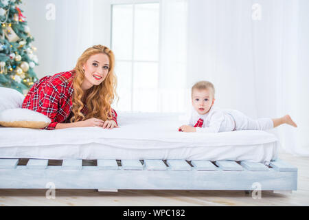 Mutter und Sohn in karierten Schlafanzug sitzen auf dem Bett in der Nähe des Neuen Jahres Baum und lächelnd Stockfoto