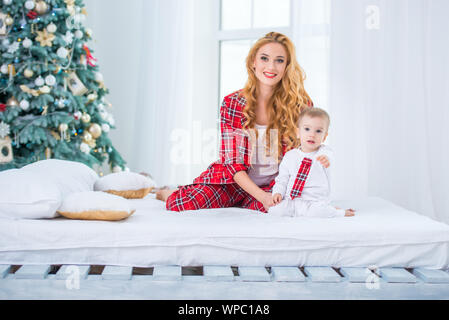 Mutter und Sohn in karierten Schlafanzug sitzen auf dem Bett in der Nähe des Neuen Jahres Baum und lächelnd Stockfoto