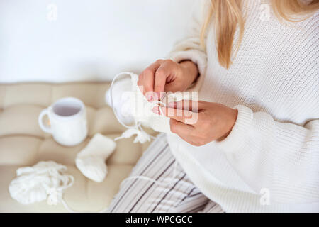 Blonde Mädchen sitzt auf einem weichen Kissen in einem weißen Pullover und Strick Baby Socken aus Wolle Stockfoto