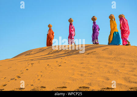 Jaisalmer, Rajasthan, Indien - 18. April 2018: Frauen, die schwere Krüge Wasser auf den Kopf und Fuß auf gelbem Sand dune im heißen Sommer d Stockfoto