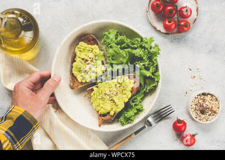 Toast mit Avocado, Samen, serviert mit grünem Salat. Ansicht von oben. Männliche Hand hält Platte mit vegan vegetarisch essen. Tabelle Ansicht von oben. Konzept der sauberen eati Stockfoto