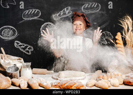 Süße Junge in Schürze und Hut werfen Mehl, Pulver in der Luft Spaß beim Teig machen. Stockfoto