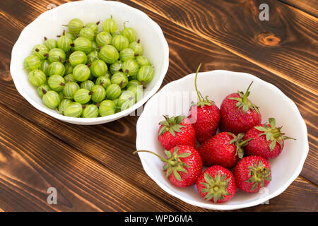 Roten saftigen sonnengereiften Erdbeeren und grüne Stachelbeeren in weißen Schüsseln auf dunklen Holztisch. Flach. Stockfoto