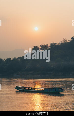 Die traditionellen laotischen Holz langsam Boot auf dem Mekong River in der Nähe von Luang Prabang, Laos bei Sonnenuntergang. Goldene Stunde, Sonne im Wasser widerspiegelt Stockfoto