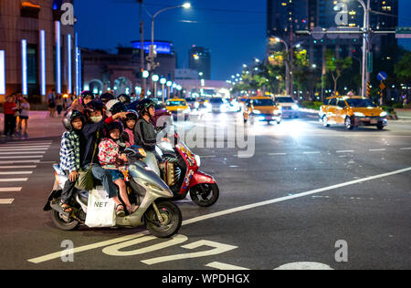 Taiwan: Mutter mit zwei Kindern auf einem Roller zeigt auf etwas außerhalb des Rahmens. Die Menschen warten auf Motorroller an einer Ampel in der Nacht Stockfoto