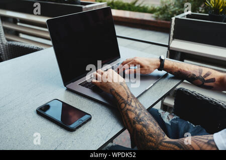 Jungen tätowierten Mann bei der Arbeit am Notebook im Cafe. Zurück Ansicht von der menschlichen Hände mit Tattoo mit Laptop an coworking Büro Schreibtisch. Hübscher Kerl mit der Texteingabe auf Stockfoto