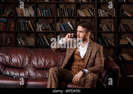 Junge Bartgeier Geschäftsmann in einem braunen Anzug im Gespräch am Telefon mit einem Lächeln auf seinem Gesicht, während in der Bibliothek Büro zu sitzen. Stockfoto