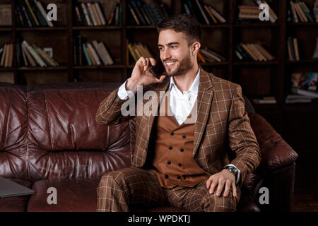 Junge Bartgeier Geschäftsmann in einem braunen Anzug im Gespräch am Telefon mit einem Lächeln auf seinem Gesicht, während in der Bibliothek Büro zu sitzen. Stockfoto