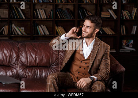 Junge Bartgeier Geschäftsmann in einem braunen Anzug im Gespräch am Telefon mit einem Lächeln auf seinem Gesicht, während in der Bibliothek Büro zu sitzen. Stockfoto