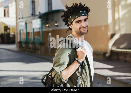 Die Jungen gut aussehenden bärtigen Mann mit Dreadlocks Frisur bei Camera suchen, Outdoor hipster Portrait auf der Herbsttagung des Europäischen Straße. Touristen Backpacker, st Stockfoto