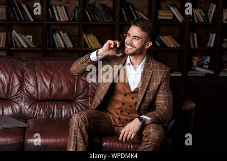 Junge Bartgeier Geschäftsmann in einem braunen Anzug im Gespräch am Telefon mit einem Lächeln auf seinem Gesicht, während in der Bibliothek Büro zu sitzen. Stockfoto